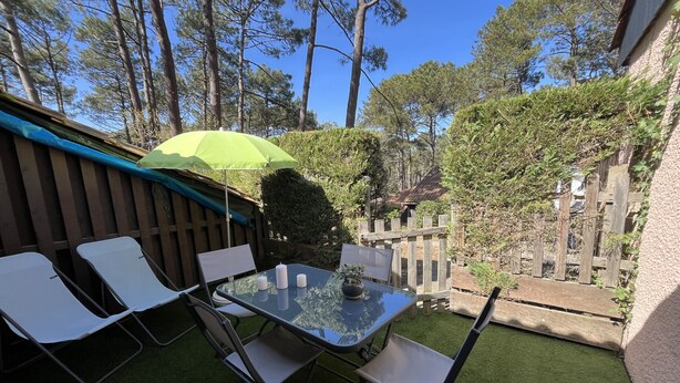 A patio with a glass table, two chairs, a green umbrella, and a wooden fence. - Maison cocon by Agence Bru - 684414 (Lacanau)