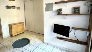 A room with a wooden cabinet, a small round table, a mounted TV, and shelves with decorative items. - Maison cocon by Agence Bru - 684414 (Lacanau)