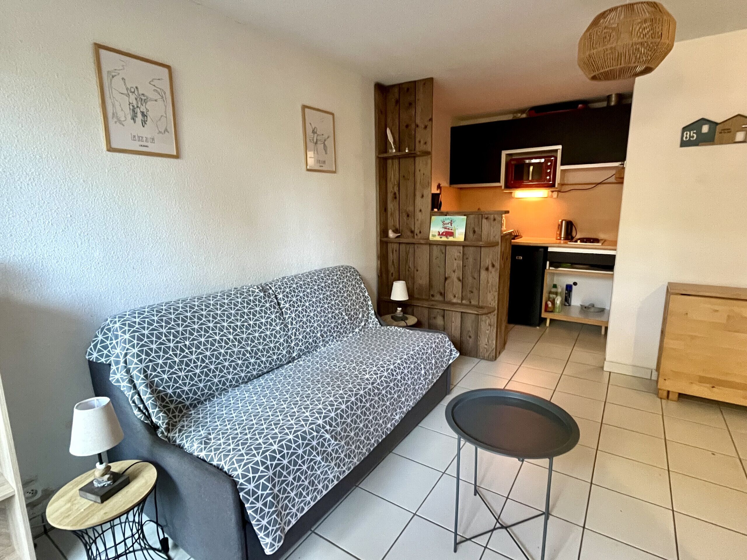 Ein gemütlicher Wohnbereich mit einem gemusterten Sofa, einem kleinen runden Tisch mit Lampe und einer Küchenzeile im Hintergrund.