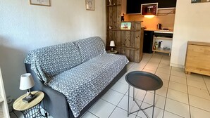 A cozy living area with a patterned sofa, a small round table with a lamp, and a kitchenette in the background. - Maison cocon by Agence Bru - 684414 (Lacanau)