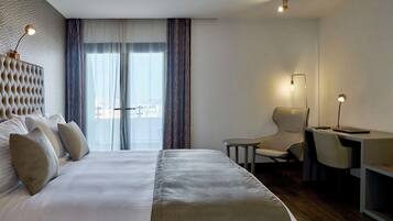 Et moderne hotelværelse med en stor seng, et skrivebord og en stol. Der er et vindue med gardiner, en lampe og udsigt over byen.