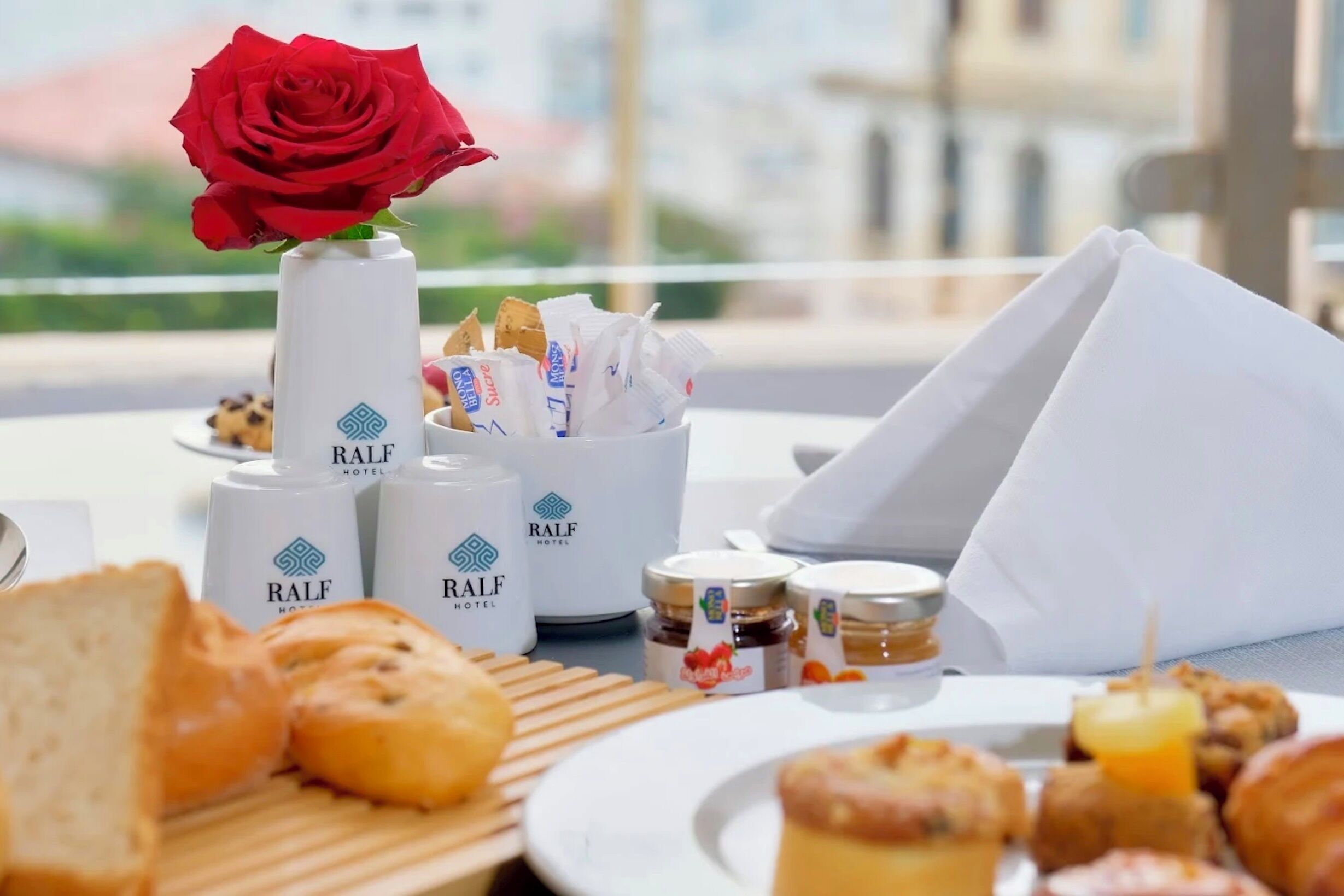 A breakfast setting with bread, pastries, condiments, and a red rose on a table.