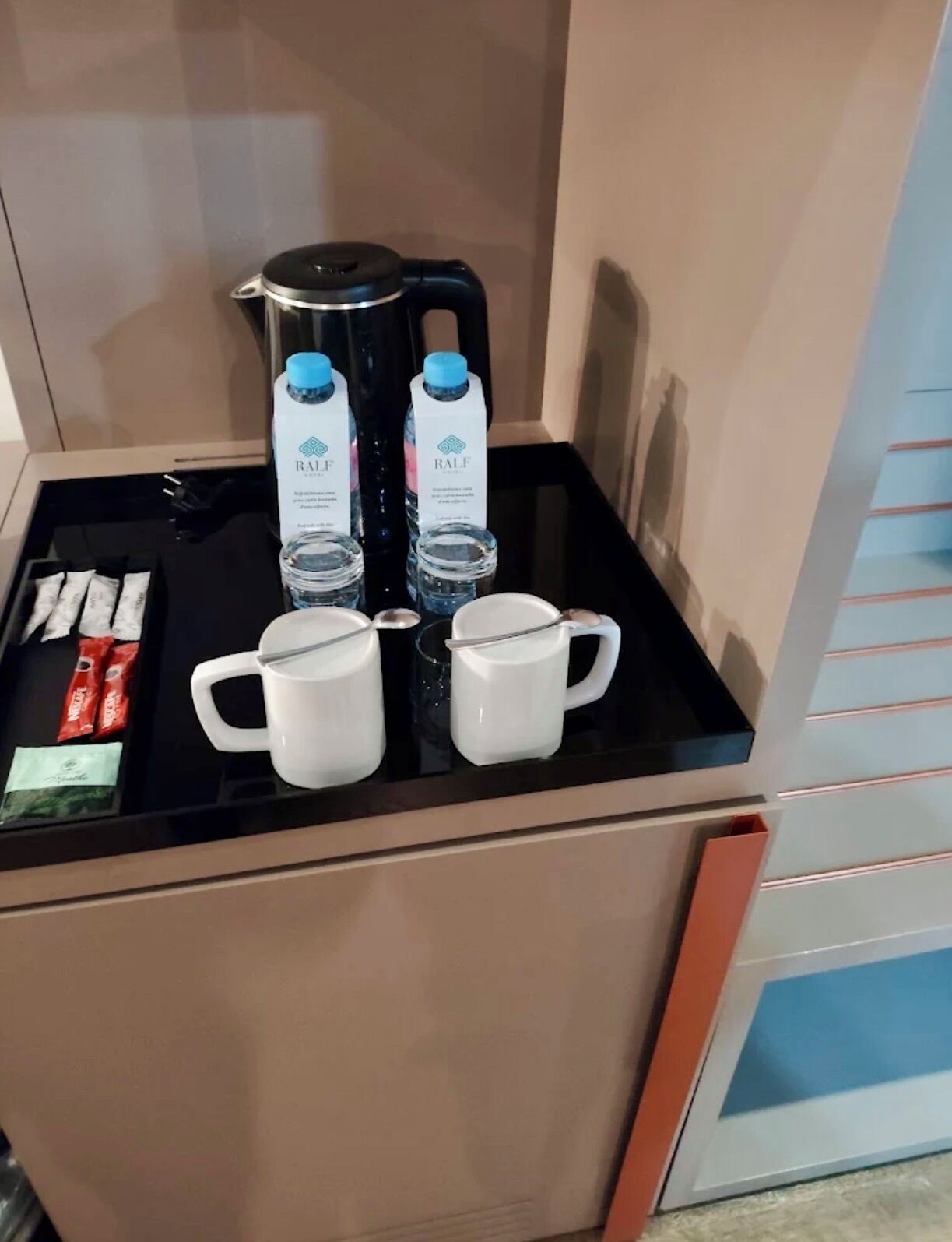 A minibar with a coffee maker, two water bottles, two white mugs, and some snacks.
