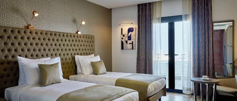Ein Hotelzimmer mit zwei Betten, einem gepolsterten Kopfteil, einem kleinen Tisch und Blick auf die Stadt durch große Fenster.