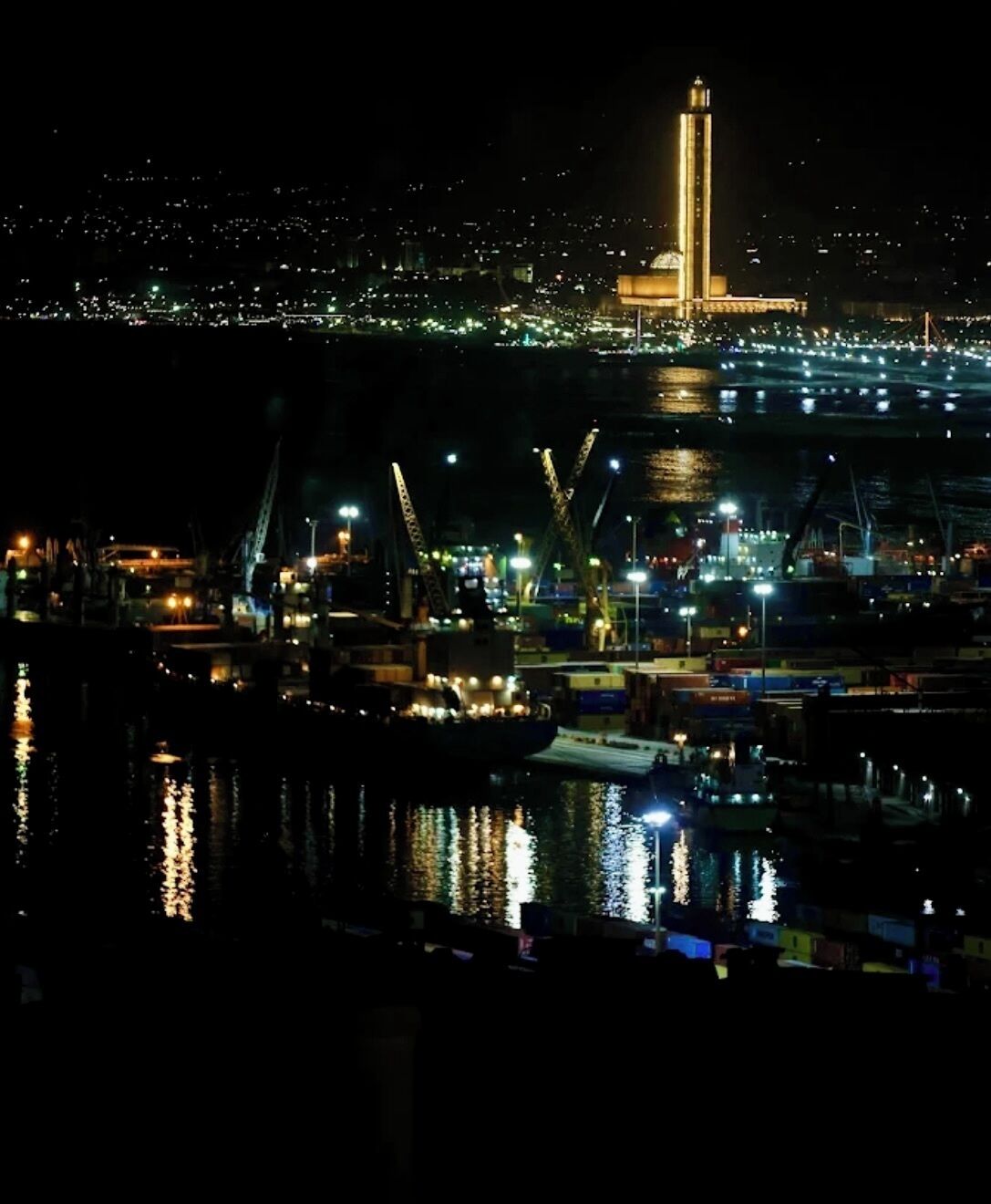 A nighttime view of a harbor with a prominent tower and a mosque.