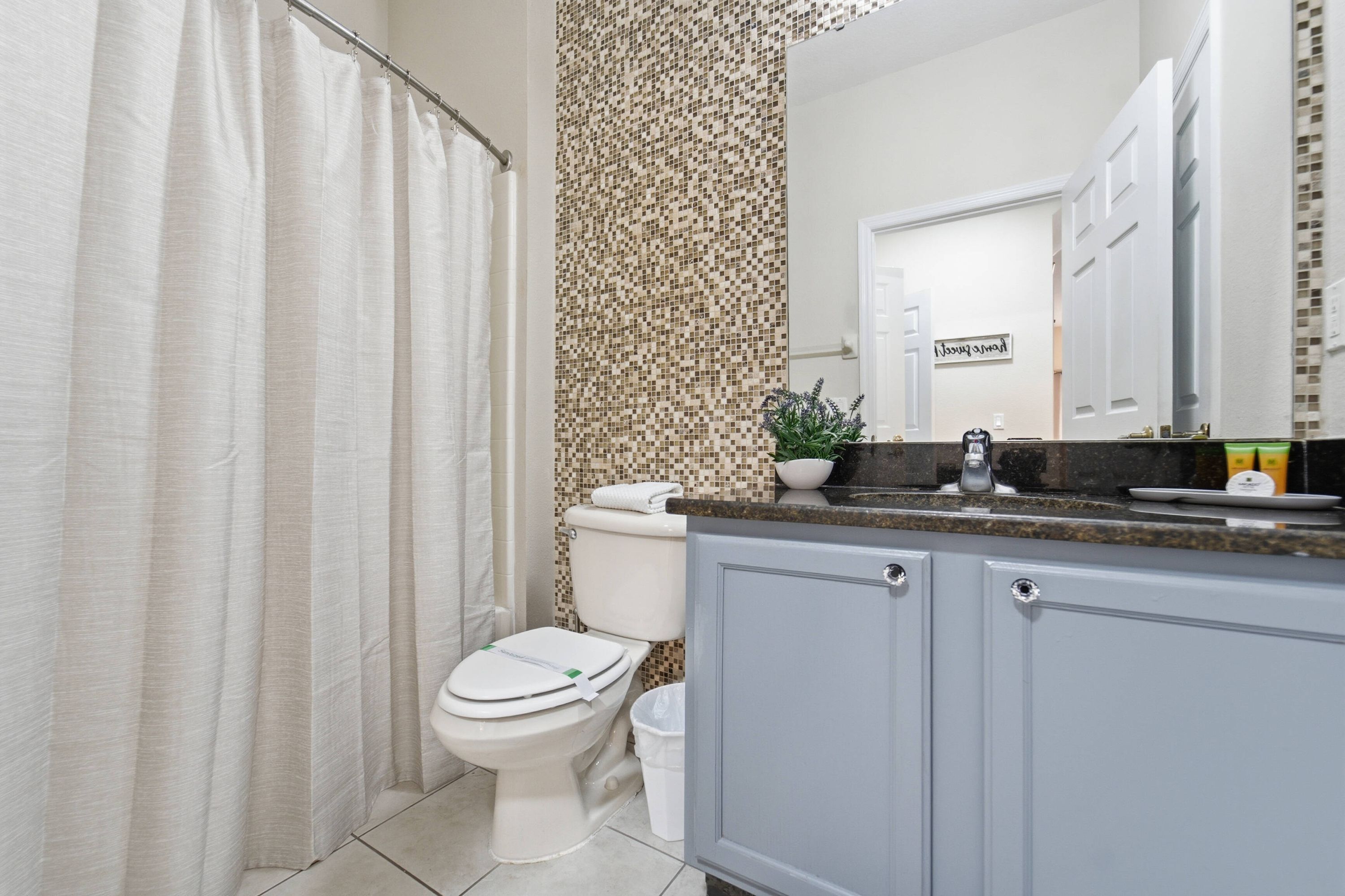 A bathroom with a toilet, a shower curtain, a sink with a mirror, and a tiled backsplash.