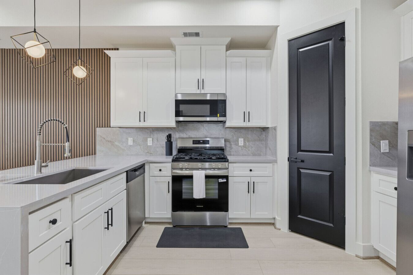 A modern kitchen with white cabinets, a black door, stainless steel appliances, and pendant lighting.