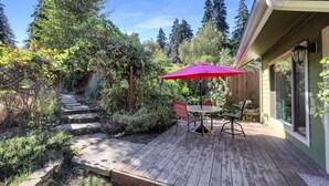 A wooden deck with a red umbrella, a table, and chairs, surrounded by greenery and a stone pathway. - Peaceful Garden Cottage Near UofO (Eugene)