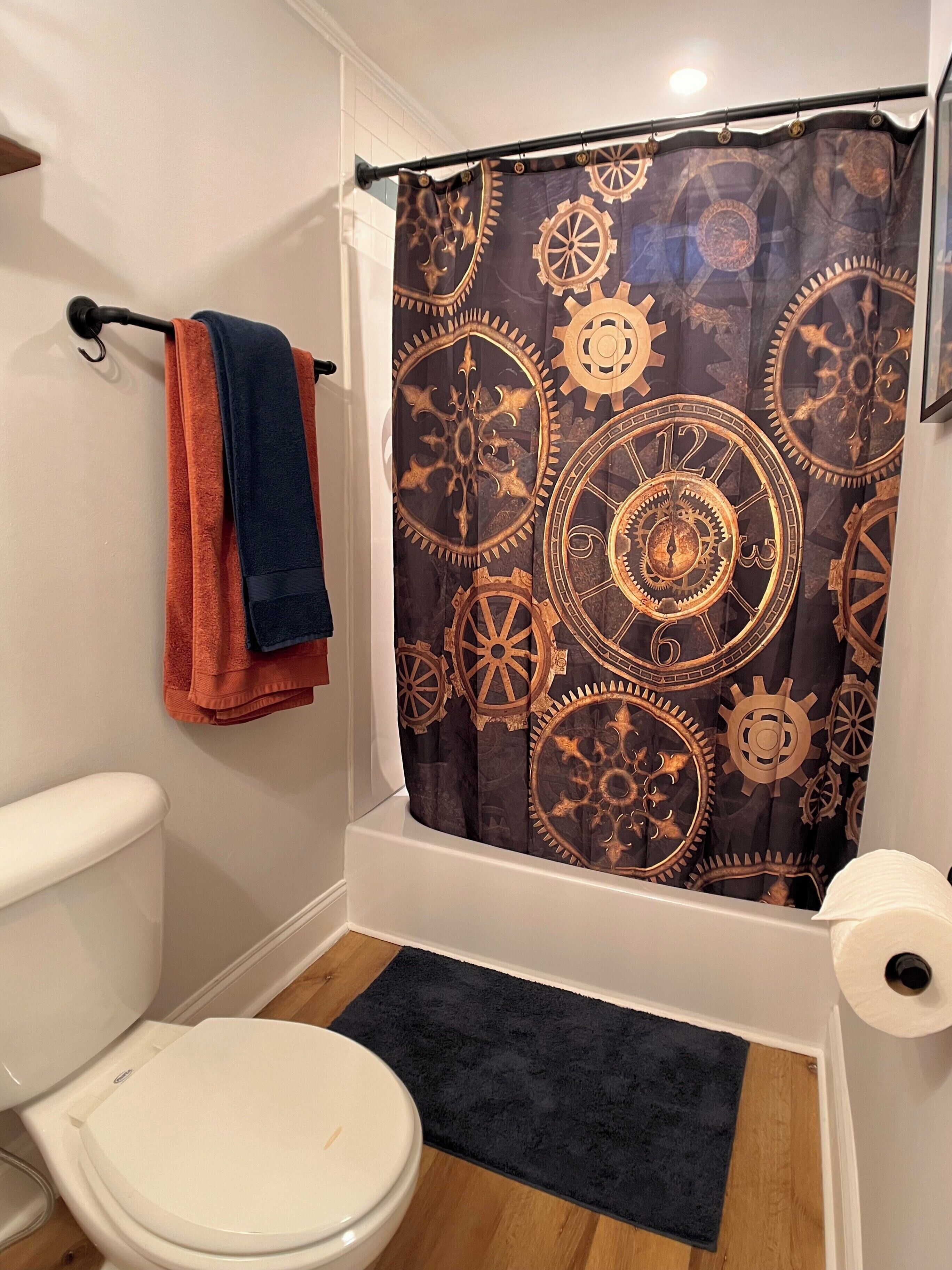 A bathroom with a white toilet, a dark blue shower curtain with gear patterns, and a pair of orange and blue towels on a rack.