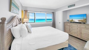 Ein Schlafzimmer mit einem großen Bett, einem Nachttisch, einer Kommode und einem Fernseher. Es gibt ein Fenster mit Blick aufs Meer.