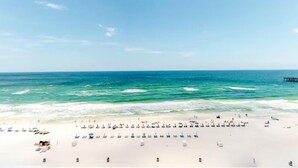 A beach with rows of sun loungers and umbrellas, a pier in the background, and calm sea waves. - 🏖️Beachfront | Gulf Views | 2BR Sterling Reef 505🏖️ (Panama City Beach)