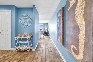 Un couloir aux murs bleus, une porte blanche, un miroir décoratif, une étagère en métal avec des bougies et un grand tableau abstrait.