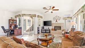 A spacious living room with a large brown sofa, a glass coffee table, a ceiling fan, a flat-screen TV, and a fireplace. - Golf-View Home | Indian Palms Resort Pool & Patio (Indio)