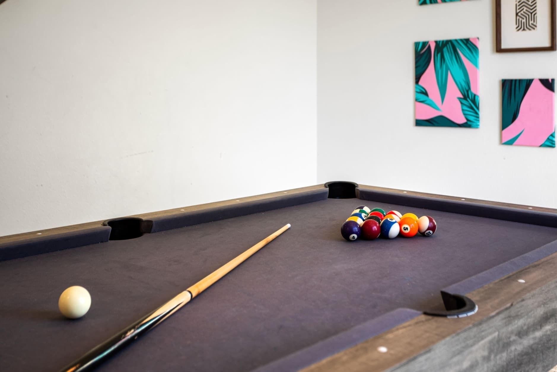 Une table de billard avec des billes et une queue, un mur blanc orné de tableaux encadrés et un plancher en bois.
