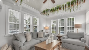 A living room with a grey sectional sofa, a wooden coffee table, and a lamp. There are white windows with blinds, a ceiling fan, and hanging plants. - La Barra #101 (St. Pete Beach)