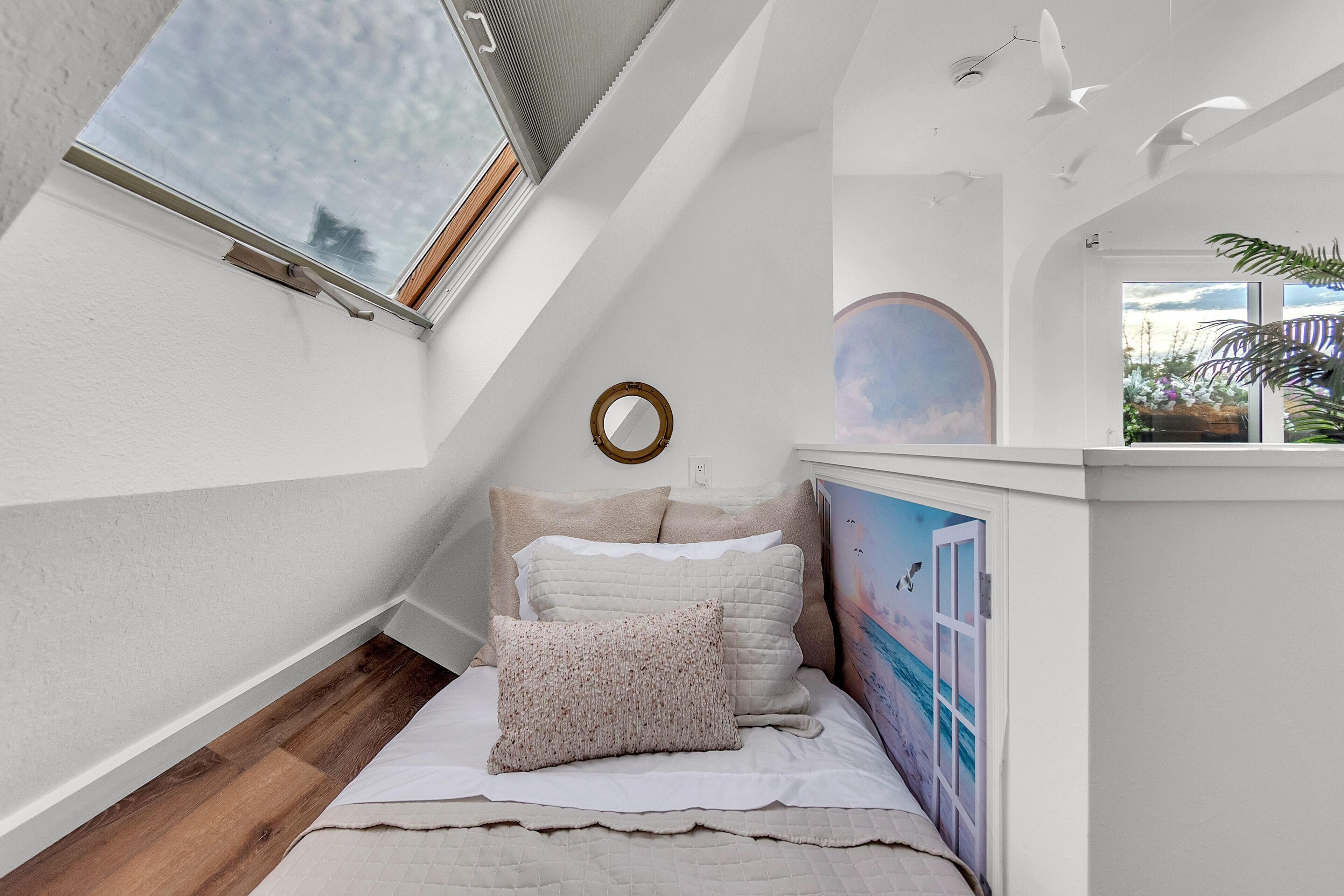 Een slaapkamer met een schuine kap, een dakraam, een bed met veel kussens, een spiegel aan de muur en een muurschildering van een strand.