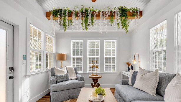 Een woonkamer met een grijze bank, een gestreept fauteuil, een houten salontafel met kaarsen en een plant, en een hangende plantenarrangement boven de ramen.