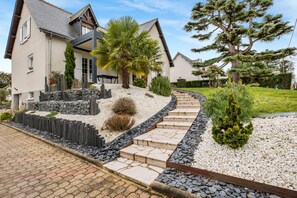 Une maison moderne avec un chemin de pierre menant à l’entrée, entourée de jardins bien entretenus et d’un grand arbre.