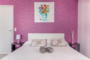 Ein Schlafzimmer mit Bett, Kissen, Nachttisch, Lampe, einer Vase und einem Gemälde. Eine Tür ist ebenfalls zu sehen.