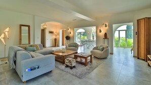 Un vaste salon comprenant un canapé, une table basse et des fauteuils, un coin repas avec une table et des chaises, et de grandes fenêtres offrant une vue sur la verdure.