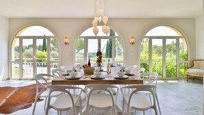 Un espace repas avec une table dressée pour six personnes, entourée de chaises blanches, et de grandes fenêtres en arc offrant une vue sur la verdure.