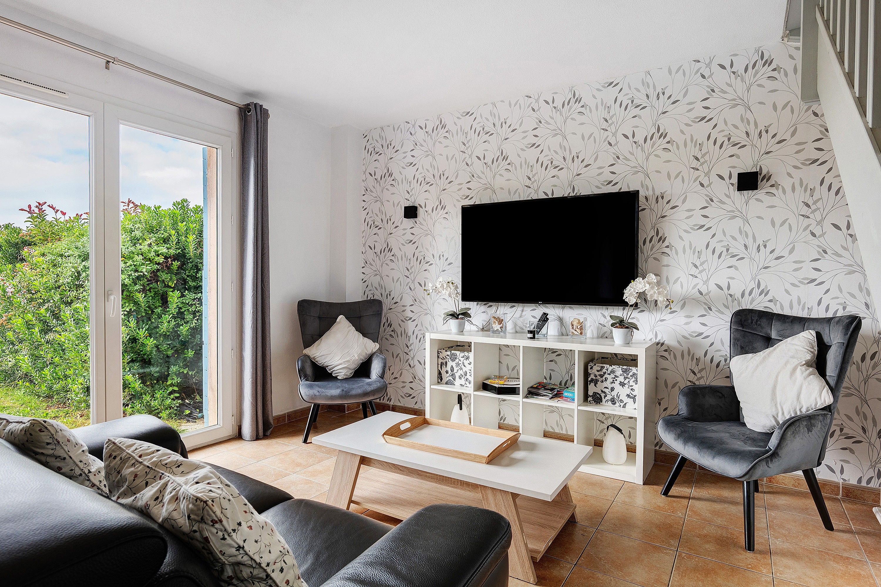 Una sala de estar moderna con un sofá, sillones, una mesa de centro y un televisor en una pared con papel tapiz con estampado de hojas.