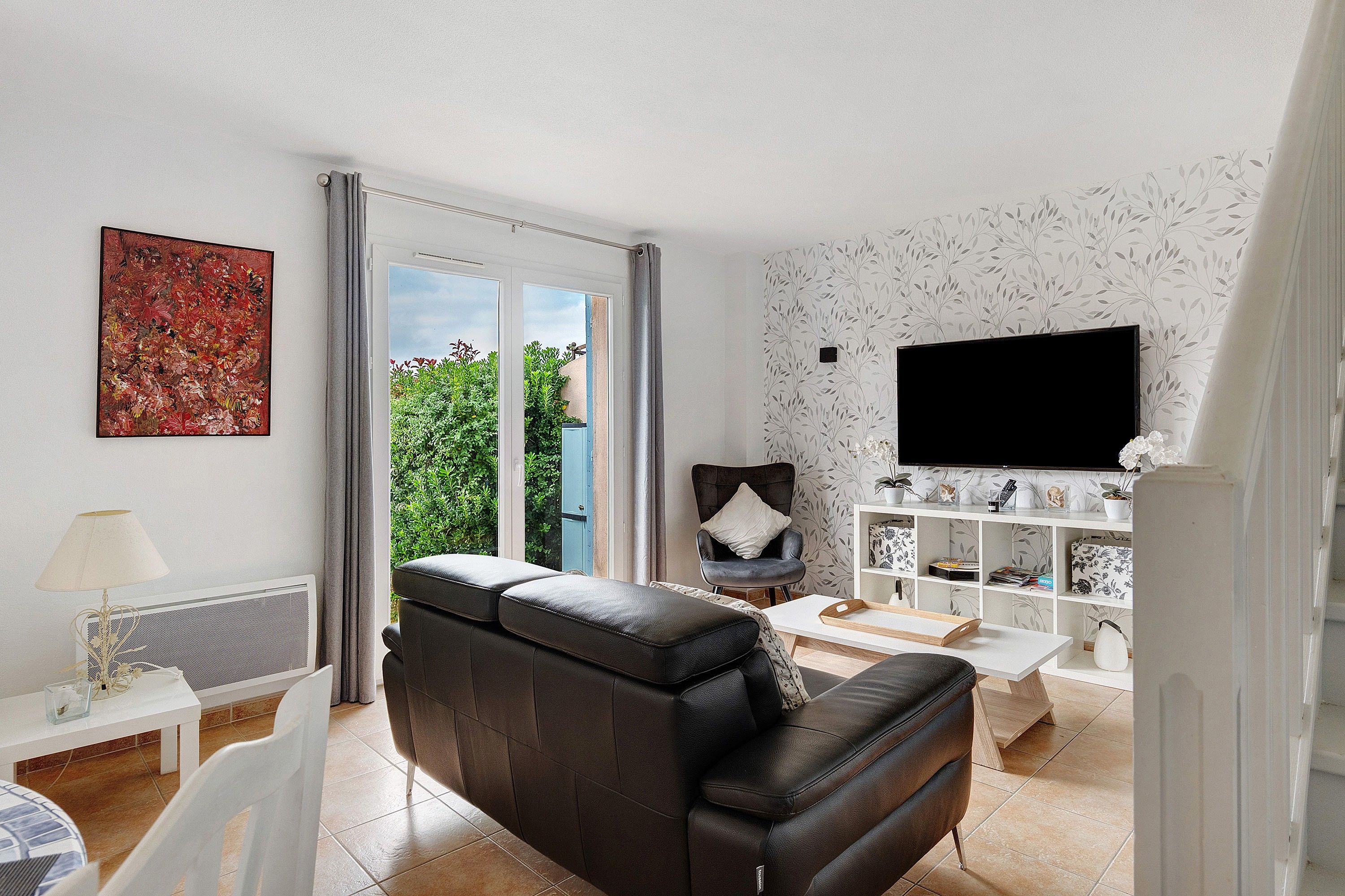 Una sala de estar con un sofá de cuero negro, una mesa de centro blanca, un televisor montado en la pared y una ventana con cortinas.