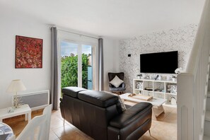 A living room with a black leather sofa, a white coffee table, a TV mounted on the wall, and a window with curtains. - Les Villas du Port Minervois - A6 (Homps)