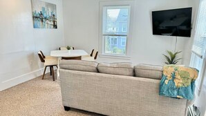 A living room with a sofa, a dining table with chairs, a flat-screen TV mounted on the wall, and a window with blinds. - Private Downtown Bar Harbor Four-Apartment Home (Bar Harbor)