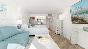 Ein modernes, gut beleuchtetes Zimmer mit einem hellblauen Sofa, einem weißen Couchtisch und einem Bett mit weißer Bettwäsche. Im Hintergrund ist ein Küchenbereich mit weißen Schränken und einer Mikrowelle erkennbar.