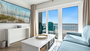 Ein Zimmer mit einem großen Fenster, einem Sofa, einem weißen Tisch und einem Gemälde mit einer Strandansicht.
