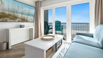 A room with a large window, a sofa, a white table, and a painting of a beach scene.