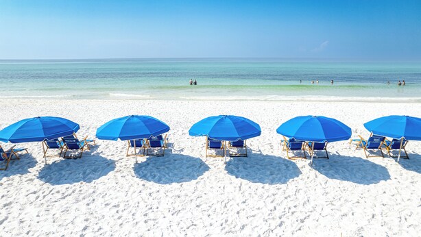 Ein Strand mit fünf blauen Sonnenschirmen und Liegestühlen, die in einer Reihe aufgereiht sind.