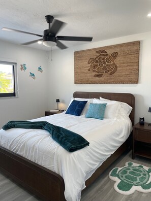 A neatly made bed with white bedding and a teal throw blanket, a ceiling fan, a wall with a turtle artwork, and a turtle rug on the floor. - Key Colony Beach Home Casa Sunny Sea Key Colony Beach, Fl Keys (Key Colony Beach)