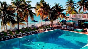 A swimming pool with lounge chairs and umbrellas, surrounded by palm trees and a view of the ocean.