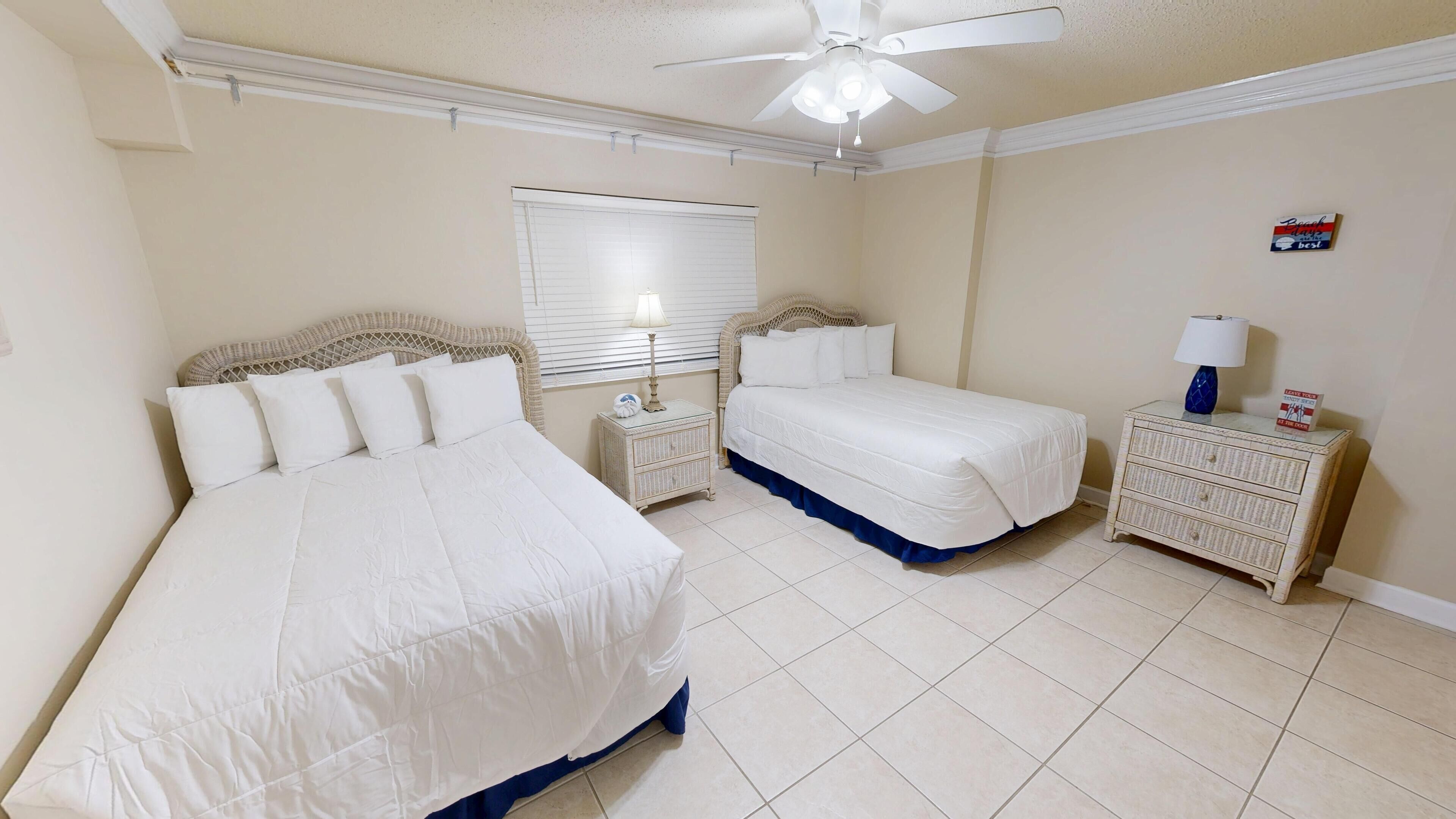 Quarto com duas camas, ventoinha no teto, janela com persianas e mesa de cabeceira com abajur.