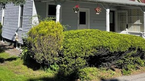 Una casa de dos plantas con techo a dos aguas, un porche con puerta blanca y un pequeño jardín con arbustos.
