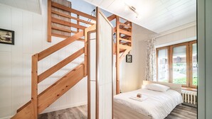 A bedroom with a wooden staircase, a bed with a white mattress, and a window with a view of greenery.