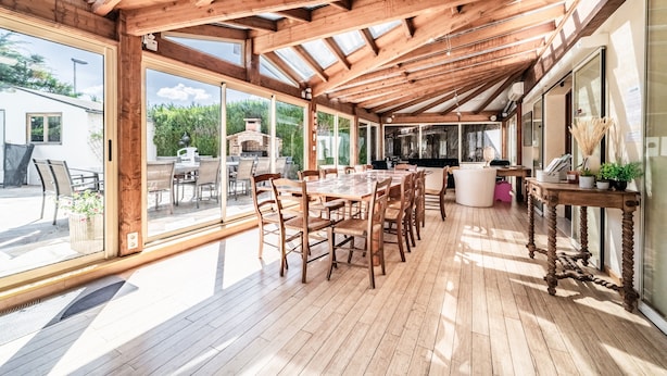 A covered patio with a wooden table and chairs, large windows, and a view of the outdoors.