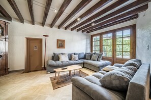 A living room with a sectional sofa, a wooden coffee table, and a large window with a view of greenery. - Clos Léonard – Country House with Pool for 14 Guests near Annecy (La Balme-de-Sillingy)