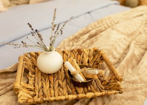 Una cesta tejida con un jarrón blanco que contiene lavanda seca y un pequeño cuenco con un objeto enrollado sobre una superficie de tela con textura.