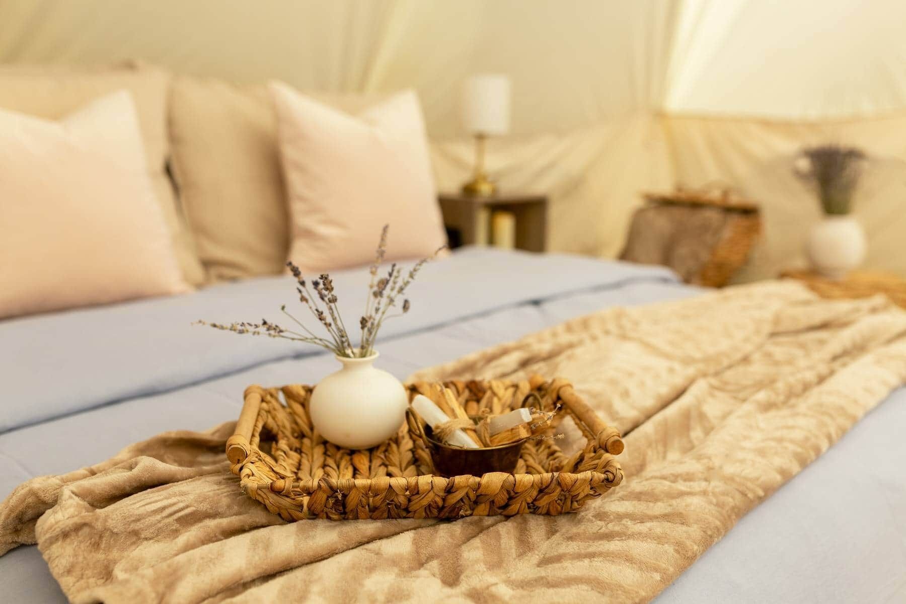 A bed with a beige blanket, a woven tray with a white vase and dried herbs, and a small bowl of cinnamon sticks.