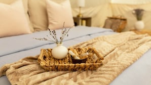 Un lit recouvert d’une couverture beige, un plateau en osier avec un vase blanc et des herbes séchées, et un petit bol rempli de bâtons de cannelle.