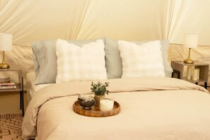 Un lit avec une couette beige, deux oreillers, une table de chevet avec une lampe et un plateau sur lequel se trouvent une bougie et une plante.