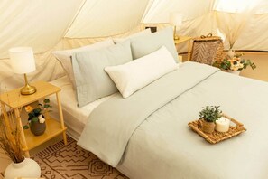 Alojamiento en tienda de campaña con cama, lámparas de noche, mesita de noche con lámpara, planta pequeña y cesta decorativa.