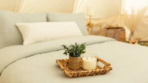 Ein Bett mit einer hellen Decke, ein kleiner gewebter Korb mit Kerze und Pflanze, und ein weißes Kopfkissen.