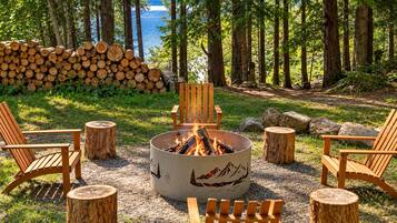 A fire pit with chairs and logs in a forested area with a lake view.