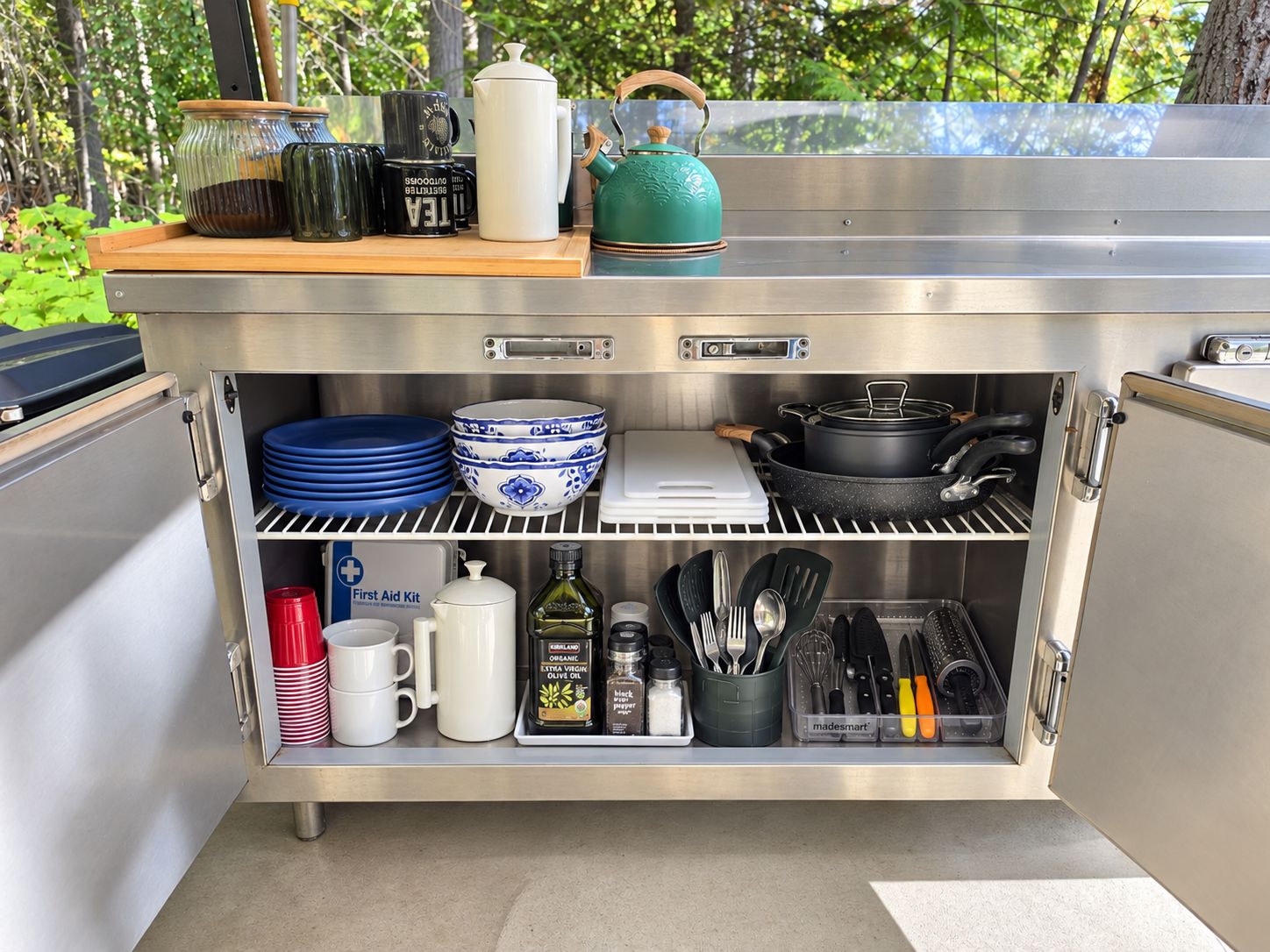 A stainless steel outdoor kitchen with a built-in storage unit, including a first aid kit, cooking utensils, and a kettle.