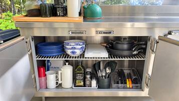 A stainless steel outdoor kitchen with a built-in storage unit, including a first aid kit, cooking utensils, and a kettle.