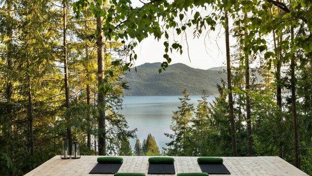 Een houten terras met yogamatten en een glazen lantaarn, met uitzicht op een meer en bos.
